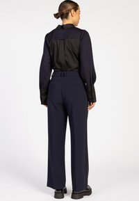 Vrouw die met haar rug naar de camera staat, gekleed in een lange mouwen top in zwart en marineblauw met brede manchetten, en bijpassende marineblauwe broek met zwarte schoenen.