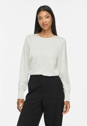 Femme aux longs cheveux foncés portant un chemisier blanc à manches longues et un pantalon noir taille haute, debout devant un fond blanc uni.