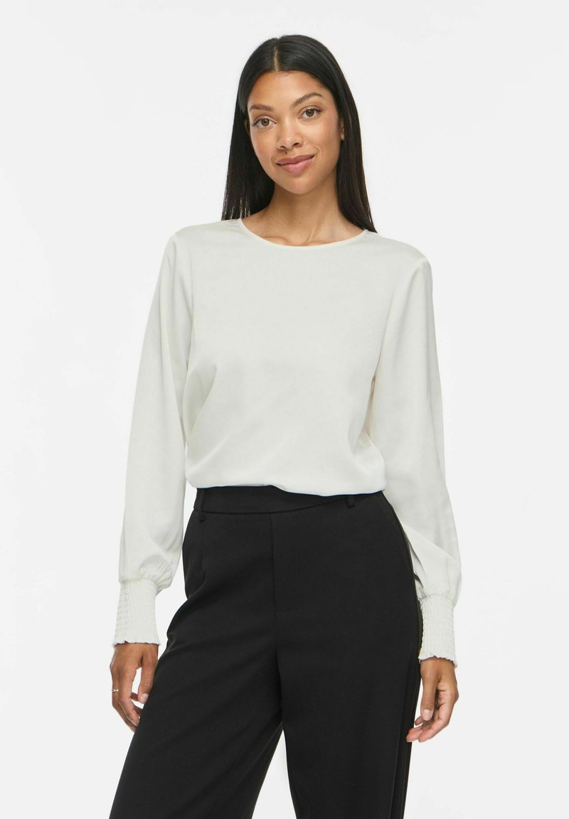 Femme aux longs cheveux foncés portant un chemisier blanc à manches longues et un pantalon noir taille haute, debout devant un fond blanc uni.
