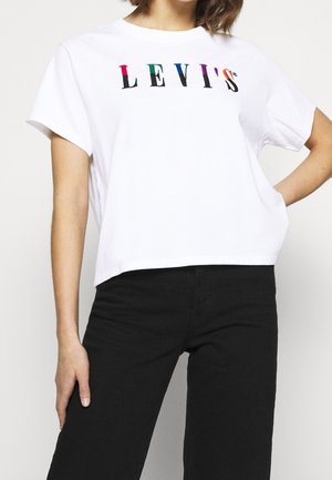 Person iført en hvid Levi's t-shirt med flerfarvede logobogstaver og sorte bukser, stående med den ene hånd på hoften.