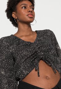 Blusa cropped preta com padrão salpicado branco, decote em V e detalhe de cordão na barra. Mangas longas com textura franzida.