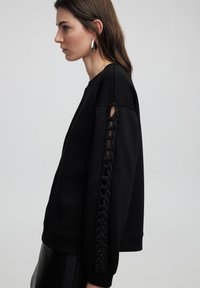 Sweatshirt noir à coupe ample, avec un détail tressé le long de la manche gauche. Tissu doux et poignets en côtes complètent le design.