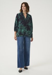 Blouse à imprimé floral bleu marine foncé avec des motifs verts, présentant un col en V et des manches trois-quarts, associée à un jean en denim bleu à jambes larges.