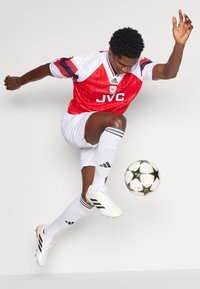 Camisa de futebol vermelha e branca, calções brancos, chutes pretos e brancos, saltando com uma bola de futebol preta e branca. Tecido texturizado com riscas.