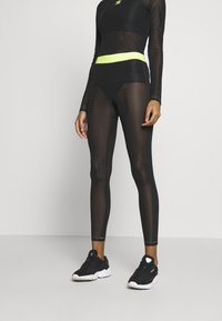 Top atletico in rete nera e leggings con cintura gialla neon, con accenti del logo e design aderente. Indossati con sneakers nere.