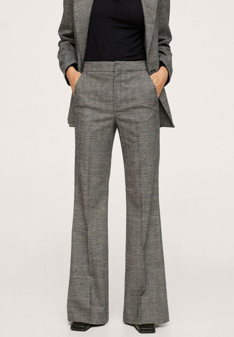 Femme portant un pantalon large à carreaux gris assorti à un blazer et un haut noir, mains dans les poches, debout sur un fond neutre.