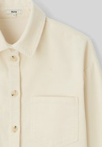 Chemise beige en velours côtelé avec un col classique, cinq boutons et une poche poitrine unique. Présente une texture côtelée subtile et une coupe décontractée.