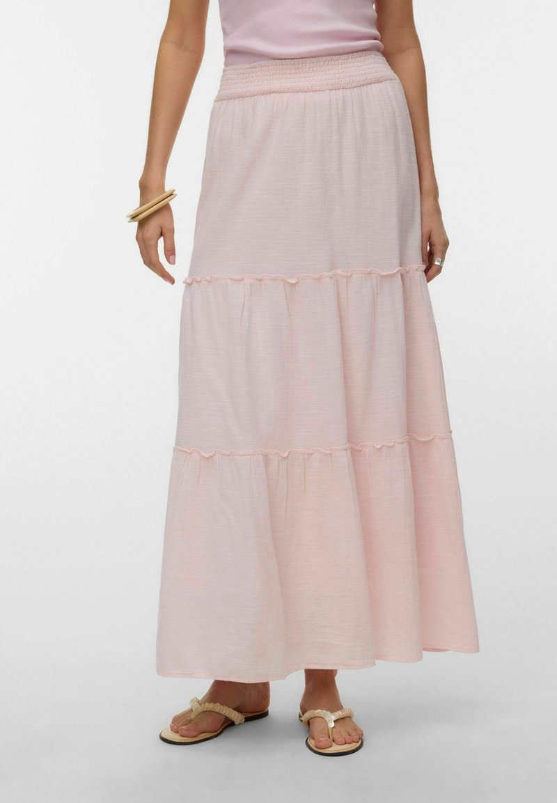 Naine kannab heledat roosat mitmekihilist maksiseelikut elastse vöökohaga, beežikaid sandaale ja puidust käevõrusid ühel randmel.