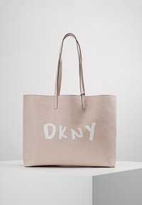 Sac cabas beige avec de longues anses reposant sur une surface blanche, logo blanc "DKNY" centré à l'avant.