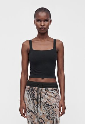 Młoda kobieta z krótkimi włosami ubrana w czarny bezrękawnik crop top oraz spodnie z abstrakcyjnym wzorem, stojąca na jasnym, jednolitym tle.