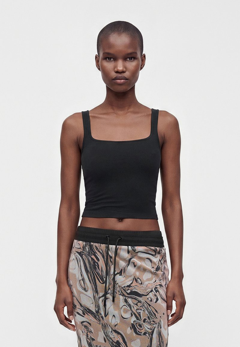 Jeune femme aux cheveux courts portant un crop top noir sans manches et un pantalon à motifs abstraits, debout devant un fond clair uni.