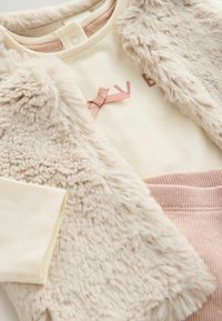 Nepbont vest in beige met een zachte textuur, een crèmekleurig shirt met een roze strik en opdruk, gecombineerd met roze geribde broeken.