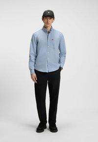 Camicia button-up blu chiaro con tasca, indossata con pantaloni neri e scarpe nere. La camicia presenta un piccolo logo rosso sulla tasca.