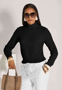 Frau in einem strukturierten schwarzen Langarmoberteil, weißen Hosen, großen weißen Ohrringen, klobigen Armbändern, Sonnenbrille und einer braunen Handtasche.