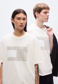 Λευκά υπερμεγέθη μπλουζάκια με ορθογώνιο γραφικό που φέρει τη φράση "TOMMY JEANS" με έντονα γράμματα. Το ύφασμα φαίνεται μαλακό, με κοντά μανίκια και άνετη εφαρμογή.