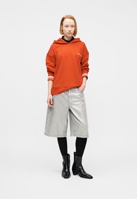 Γυναίκα όρθια, φοράει φούτερ κεραμιδί, ασημί σορτς μέχρι το γόνατο, μαύρο κολάν και μαύρα μποτάκια αστραγάλου σε λευκό φόντο.