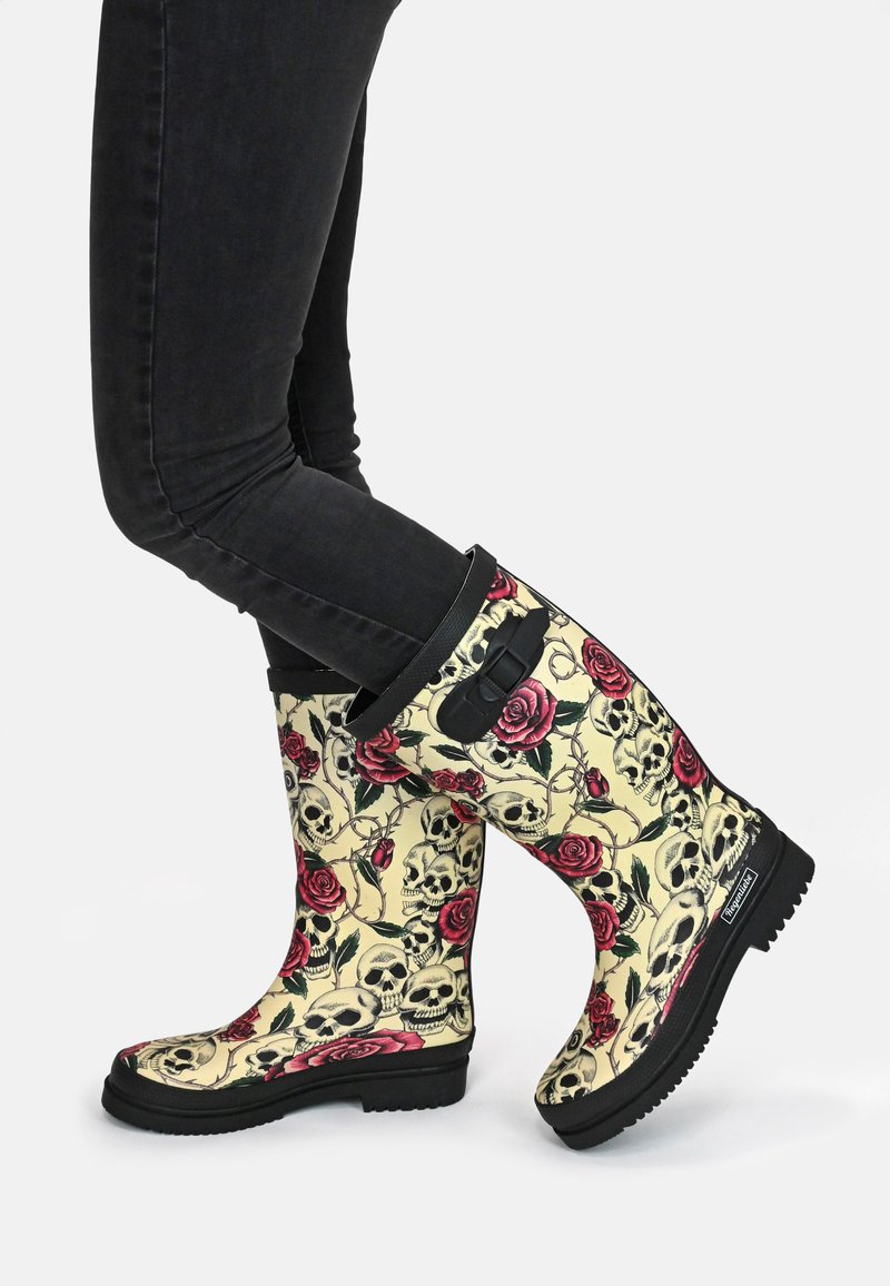 Rubber rain boots with a cream background, featuring a pattern of skulls and red roses. Black sole and top accents, pull tab for easy wear.