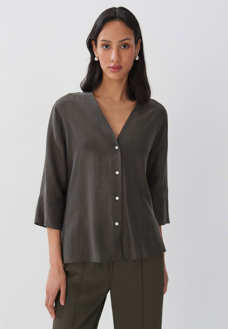 Blouse ample vert foncé avec un décolleté en V, des manches trois-quarts et des boutons blancs sur le devant. Tissu doux et texturé.