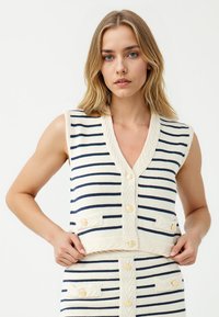 Gebreide mouwloze top met een diepe V-hals, voorzien van marineblauwe strepen op een crème achtergrond, decoratieve knopen en een textuurband in de taille.