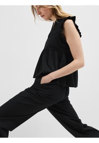 Svart ärmlös topp med volangärmar, en utsvängd design och samlad midja. Modellen bär matchande svarta byxor. Slät tygtextur.