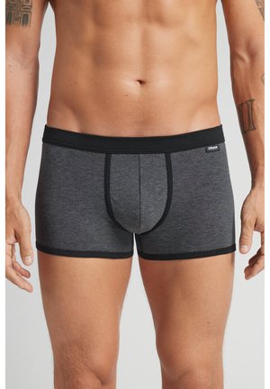 Man die zwarte en grijs gestreepte boxershorts draagt, staand met ontspannen armen langs zijn zij, waarbij de romp en bovenbenen zichtbaar zijn.