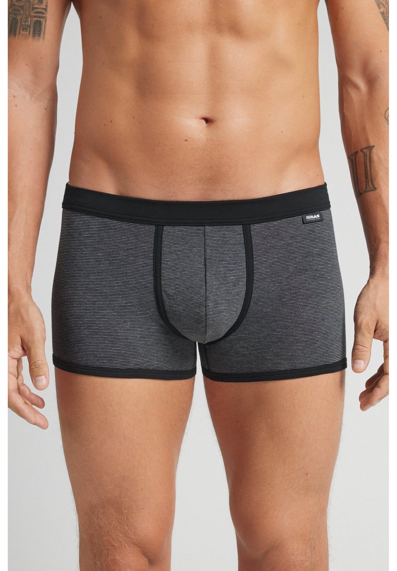 Muž stojící v černých a šedých pruhovaných boxerkách, paže má volně podél těla, vidět je trup a horní část nohou.