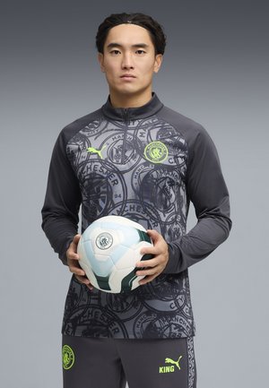 Hombre con camiseta de entrenamiento del Manchester City sostiene un balón de fútbol blanco y verde del Manchester City sobre un fondo gris.