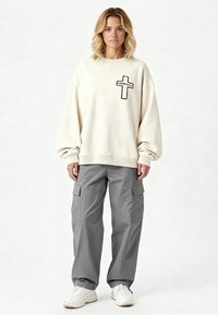 Femme blonde portant un sweat-shirt crème oversize avec l'impression croisée "Karma Kills", un pantalon cargo gris et des baskets blanches, debout devant un fond blanc.