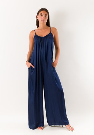 Donna in piedi con le mani nelle tasche che indossa una tuta blu navy a gamba larga e tacchi beige aperti sulle dita, su uno sfondo bianco semplice.