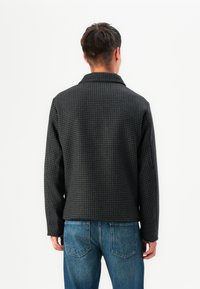 Veste à motif à carreaux noirs avec col, en tissu texturé, et coupe décontractée. Portée par-dessus un jean en denim bleu, comportant des détails au dos.