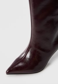 Botte à bout pointu en cuir verni marron foncé avec des coutures visibles sur le devant et les côtés, sur un fond blanc uni.