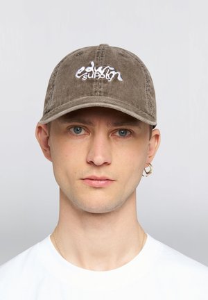 Giovane uomo dalla pelle chiara, occhi azzurri, che indossa un berretto marrone con la scritta bianca "edsvn supply", una camicia bianca e un orecchino a cerchio dorato.