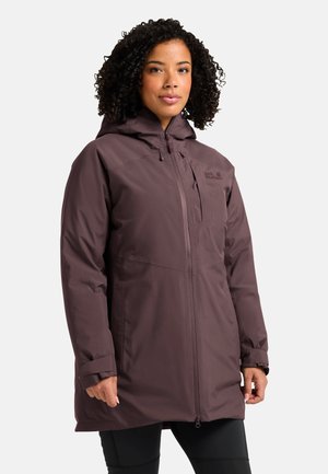 Donna con capelli ricci che indossa un lungo giubbotto invernale viola con cappuccio, zip e logo sul petto, in piedi su uno sfondo semplice.
