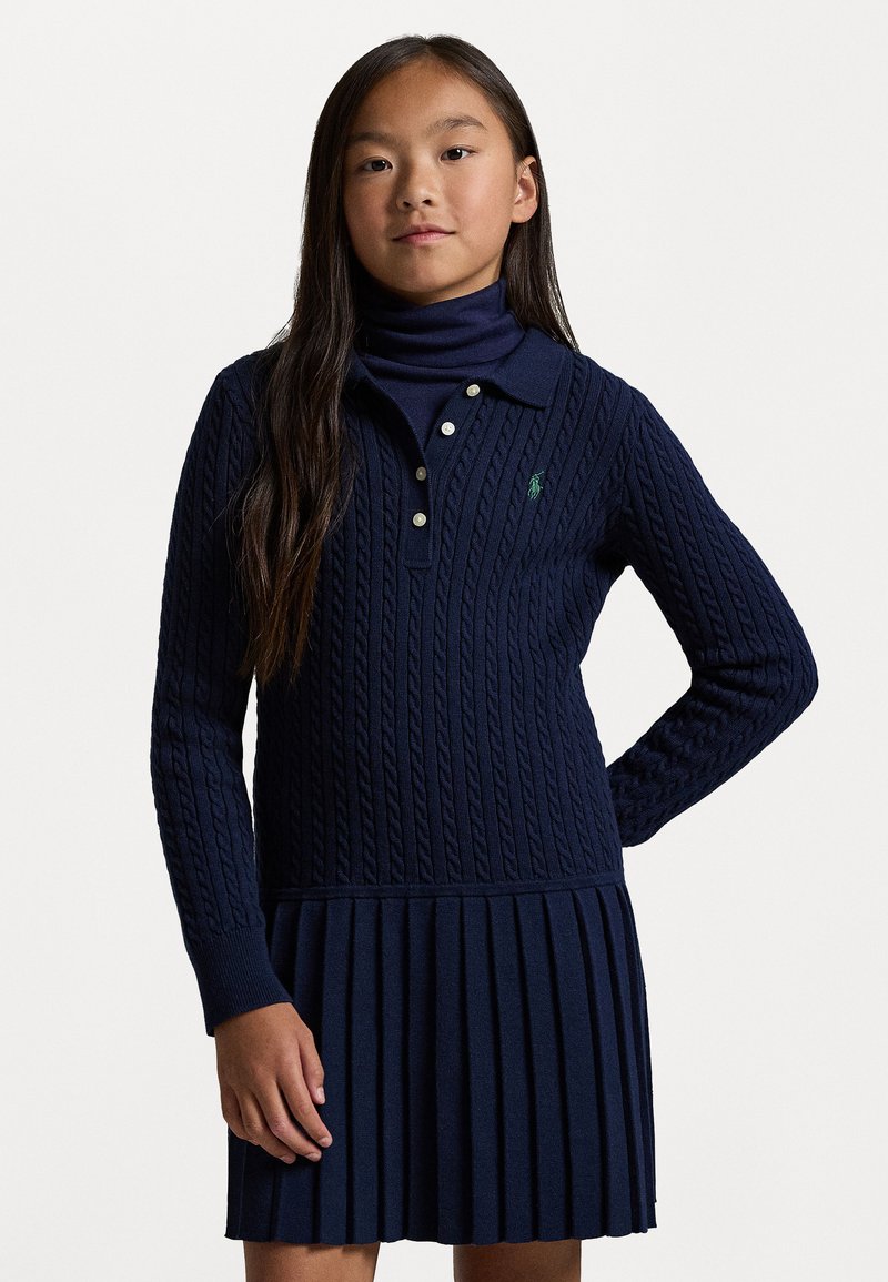 Σκούρο μπλε πλεκτό φόρεμα με καπιτονέ σχέδιο, που διαθέτει γιακά, τρία κουμπιά, μακριά μανίκια και πλισέ φούστα. Λεπτομέρεια πράσινου λογότυπου.