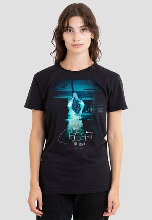 Junge Frau trägt ein schwarzes T-Shirt mit einer blauen, verzerrten Figur, die einen Baseballschläger hält, und dem Schriftzug "The Creep Tapes" auf der Vorderseite.