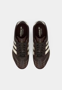 Sapatilhas desportivas de couro castanho com detalhes brancos, três riscas, sola texturizada e costura decorativa na biqueira.