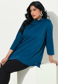 Tealfärgad tunika med hög halsringning, trekvartslånga ärmar och en avslappnad passform. Har en subtil logotyp nära fållen. Matchas med svarta byxor.