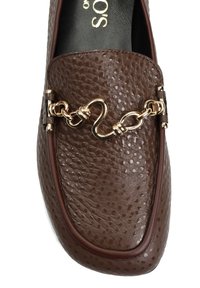 Brązowy mokasyn ze skóry o fakturze z złotym łańcuszkiem na górze oraz gładką, ciemną podeszwą wewnętrzną.