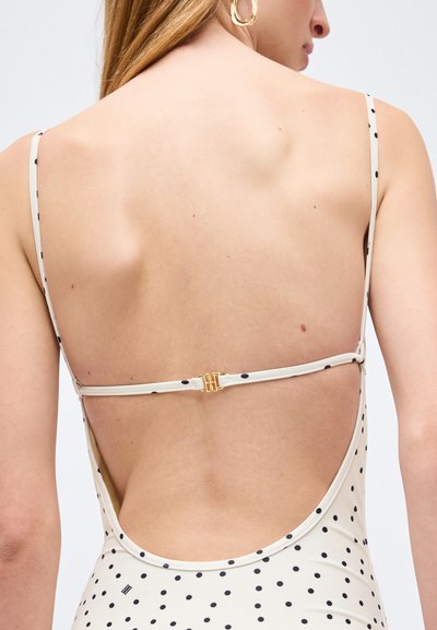 Γυναίκα που φοράει λευκό φόρεμα με πουά, λεπτές τιράντες και χαμηλή πλάτη, παρουσιάζοντας λεπτομέρεια με χρυσό κούμπωμα και μεγάλο χρυσό κρίκο στο ορατό αυτί.