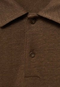 Hnědá látka polo trička s texturovaným povrchem. Obsahuje plochý límec a dva tmavé knoflíky na léze.