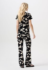 Vrouw met lang blond haar, gekleed in een bijpassende zwart-beige bloemenprint top met korte mouwen en flared broek, staande tegen een witte achtergrond.