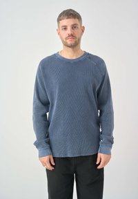 Pullover azul de tejido waffle con mangas largas, escote redondo y dobladillos con puños, combinado con pantalones negros. Textura ligera y corte relajado.