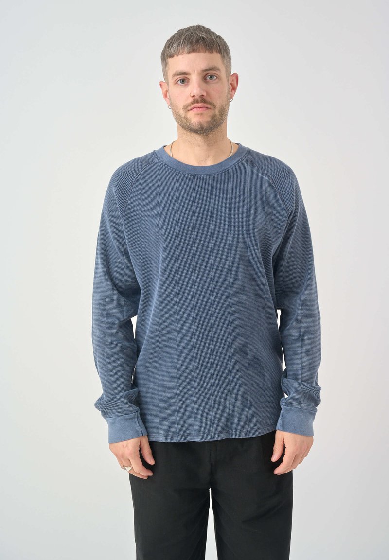 Pullover azul de tejido waffle con mangas largas, escote redondo y dobladillos con puños, combinado con pantalones negros. Textura ligera y corte relajado.