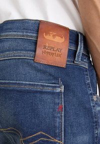 Tamno plave traperice s kožnom oznakom na struku urezanom s "REPLAY HYPERFLEX", s žutim šavovima i crvenim detaljima.