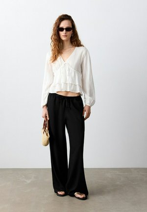 Femme aux cheveux bouclés portant des lunettes de soleil, un chemisier blanc à manches longues, un pantalon large noir, des sandales, tenant un petit sac à main tressé.