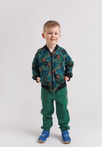 Uśmiechnięty młody chłopak stoi z rękami w kieszeniach, ubrany w zielone spodnie, niebieskie trampki i czarną kurtkę na zamek z tropikalnym wzorem liści i ananasów.