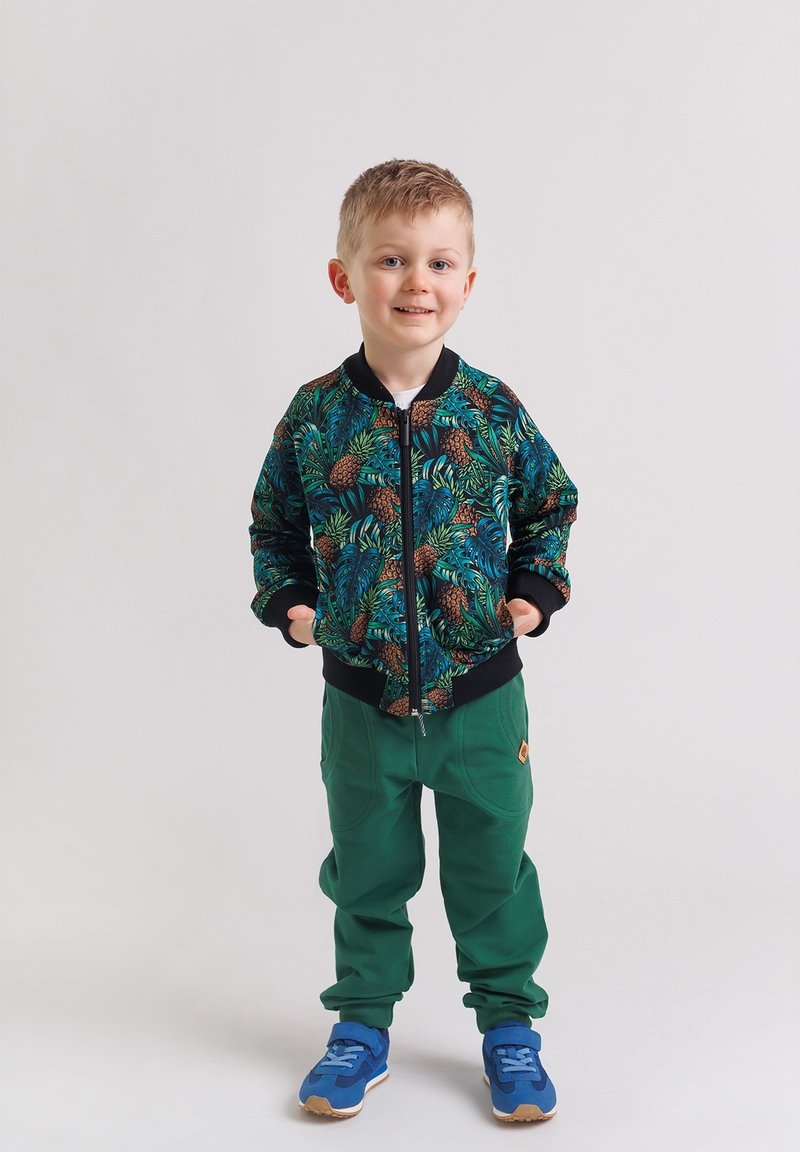 Uśmiechnięty młody chłopak stoi z rękami w kieszeniach, ubrany w zielone spodnie, niebieskie trampki i czarną kurtkę na zamek z tropikalnym wzorem liści i ananasów.