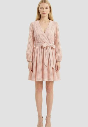 Femme portant une robe rose clair, manches longues, longueur genou, avec un motif texturé et un nœud noué à la taille, debout devant un fond uni.