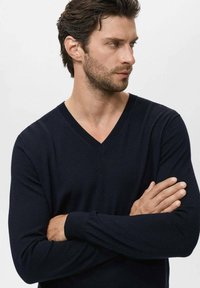 Suéter de punto suave de color azul marino con cuello en V, con mangas largas y puños acanalados, que lleva puesto un hombre con los brazos cruzados.