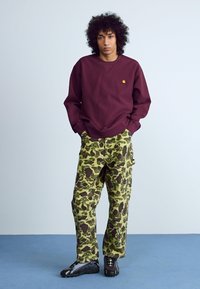 Bordeaux sweatshirt met een ronde hals, gecombineerd met groene camouflage cargo pants met bruine accenten. Zwarte sportieve schoenen maken de outfit compleet.